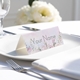 Personalised wedding place card with your name in elegant designt on folded white cardstock. Placed on a white plate with silver cutlery and a neatly folded napkin, with soft-focus white flowers in the background.