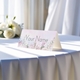 Personalised wedding place card with your name in elegant design on folded white cardstock. Displayed on a table with a flowing white tablecloth, with soft natural light and white roses in the background.