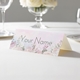 Personalised wedding place card with your name in elegant design on folded white cardstock. Displayed on a white tablecloth with wine glasses, plates, and a floral centrepiece in the softly blurred background.