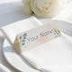 Personalised wedding place card with your name in elegant design on folded white cardstock. Positioned on a neatly folded white napkin atop a white plate, with a soft-focus white rose in the background.