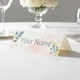 Personalised wedding place card with your name in elegant design on folded white cardstock. Displayed on a white tablecloth with wine glasses, plates, and a floral centrepiece in the softly blurred background.