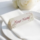 Personalised wedding place card with your name in elegant design on folded white cardstock. Positioned on a neatly folded white napkin atop a white plate, with a soft-focus white rose in the background.