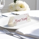 Personalised wedding place card with your name in elegant designt on folded white cardstock. Placed on a white plate with silver cutlery and a neatly folded napkin, with soft-focus white flowers in the background.