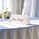 Personalised wedding place card with your name in elegant design on folded white cardstock. Displayed on a table with a flowing white tablecloth, with soft natural light and white roses in the background.