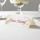 Personalised wedding place card with your name in elegant design on folded white cardstock. Displayed on a white tablecloth with wine glasses, plates, and a floral centrepiece in the softly blurred background.