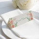 Personalised wedding place card with your name in elegant design on folded white cardstock. Positioned on a neatly folded white napkin atop a white plate, with a soft-focus white rose in the background.