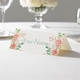 Personalised wedding place card with your name in elegant design on folded white cardstock. Displayed on a white tablecloth with wine glasses, plates, and a floral centrepiece in the softly blurred background.