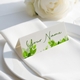 Personalised wedding place card with your name in elegant design on folded white cardstock. Positioned on a neatly folded white napkin atop a white plate, with a soft-focus white rose in the background.