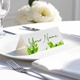 Personalised wedding place card with your name in elegant designt on folded white cardstock. Placed on a white plate with silver cutlery and a neatly folded napkin, with soft-focus white flowers in the background.