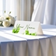 Personalised wedding place card with your name in elegant design on folded white cardstock. Displayed on a table with a flowing white tablecloth, with soft natural light and white roses in the background.