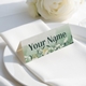 Personalised wedding place card with your name in elegant design on folded white cardstock. Positioned on a neatly folded white napkin atop a white plate, with a soft-focus white rose in the background.