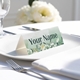 Personalised wedding place card with your name in elegant designt on folded white cardstock. Placed on a white plate with silver cutlery and a neatly folded napkin, with soft-focus white flowers in the background.