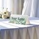 Personalised wedding place card with your name in elegant design on folded white cardstock. Displayed on a table with a flowing white tablecloth, with soft natural light and white roses in the background.