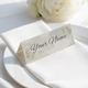 Personalised wedding place card with your name in elegant design on folded white cardstock. Positioned on a neatly folded white napkin atop a white plate, with a soft-focus white rose in the background.