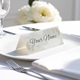 Personalised wedding place card with your name in elegant designt on folded white cardstock. Placed on a white plate with silver cutlery and a neatly folded napkin, with soft-focus white flowers in the background.