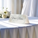 Personalised wedding place card with your name in elegant design on folded white cardstock. Displayed on a table with a flowing white tablecloth, with soft natural light and white roses in the background.