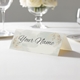 Personalised wedding place card with your name in elegant design on folded white cardstock. Displayed on a white tablecloth with wine glasses, plates, and a floral centrepiece in the softly blurred background.