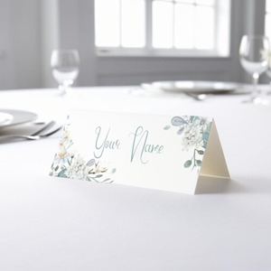 Personalised wedding place card with your name in elegant design on folded white cardstock. Placed on a table set for a formal event, with plates, cutlery, and wine glasses in the softly blurred background.