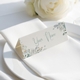 Personalised wedding place card with your name in elegant design on folded white cardstock. Positioned on a neatly folded white napkin atop a white plate, with a soft-focus white rose in the background.