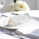 Personalised wedding place card with your name in elegant designt on folded white cardstock. Placed on a white plate with silver cutlery and a neatly folded napkin, with soft-focus white flowers in the background.