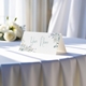 Personalised wedding place card with your name in elegant design on folded white cardstock. Displayed on a table with a flowing white tablecloth, with soft natural light and white roses in the background.