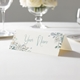 Personalised wedding place card with your name in elegant design on folded white cardstock. Displayed on a white tablecloth with wine glasses, plates, and a floral centrepiece in the softly blurred background.